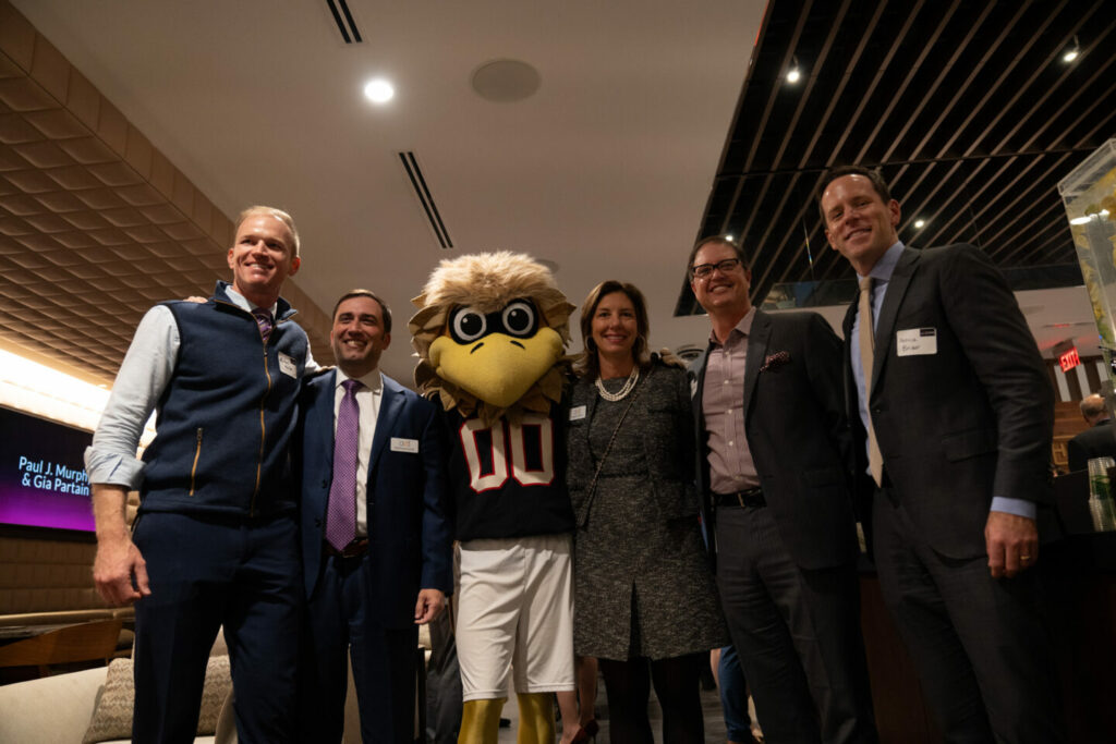 A group of four men and one woman pose with a person in a bird mascot costume wearing a sports jersey. They stand in a warmly lit indoor space, smiling for the photo.