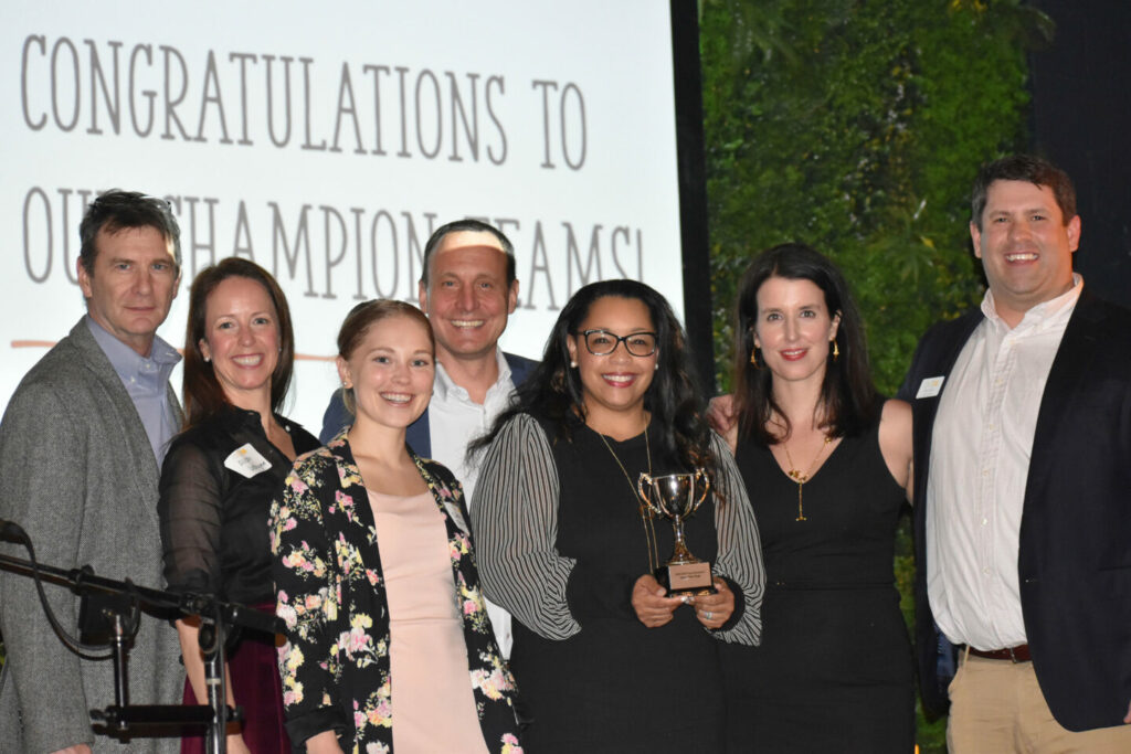 A group of seven people smiling and standing together at an event. One person in the center is holding a trophy. Behind them is a screen displaying the text "Congratulations to Our Champion Teams!.