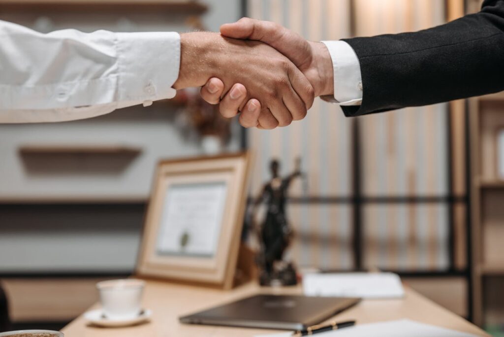 Two people shaking hands over a wooden desk in an office setting. One person wears a white shirt and the other a black suit. A laptop, a cup, and a decorative statue of Lady Justice are visible in the background.