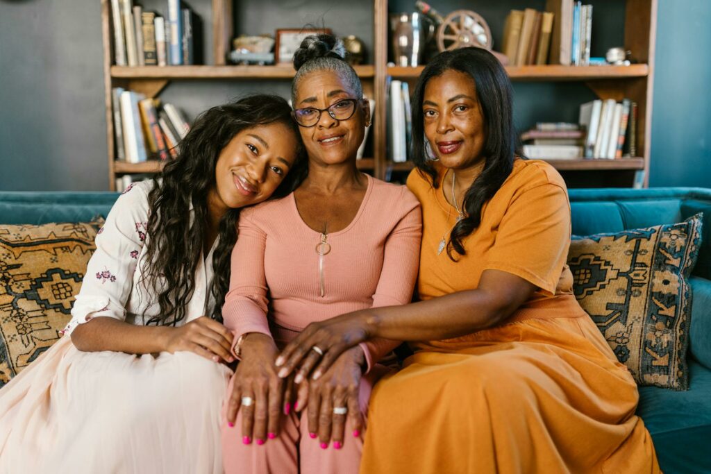 Three women sit on a sofa, smiling warmly. The woman in the middle wears glasses and a pink outfit. The woman on the left leans her head on the middle woman's shoulder, while the woman on the right wears an orange dress. A bookshelf is in the background.