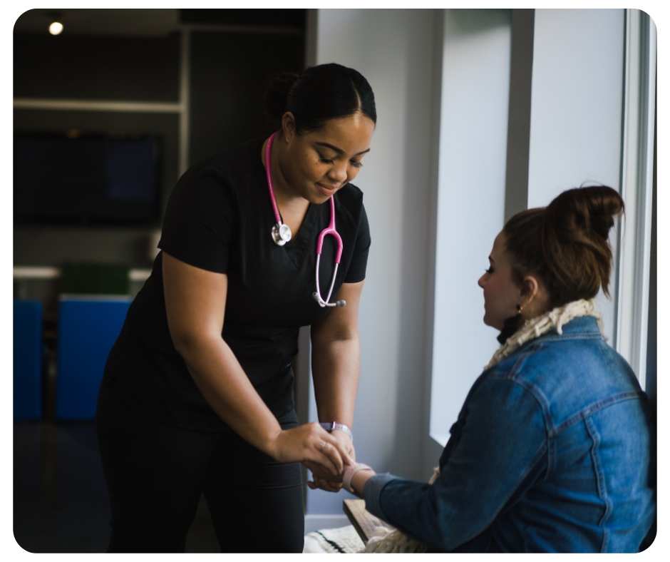 A nurse wearing black scrubs and a pink stethoscope checks the pulse of a seated woman in a denim jacket. They are in a well-lit room with large windows.