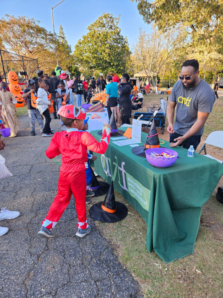 A child in a red superhero costume reaches for a piece of candy offered by a volunteer at a table with a green tablecloth. The scene is outdoors, with people in costumes, decorations, and trees in the background.