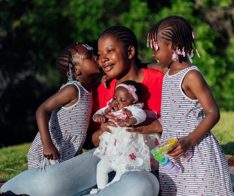 A woman sits on the grass, holding a baby, with two young girls beside her. One girl kisses her cheek, while the other holds a toy. They are all smiling and dressed in matching striped outfits. Lush greenery is in the background.