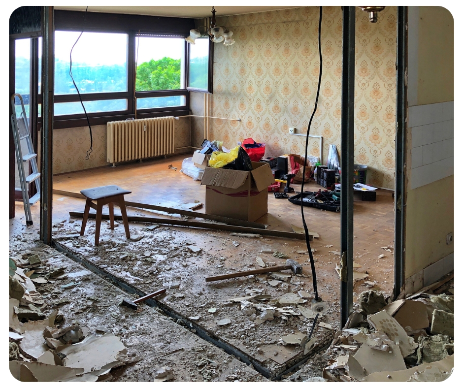 Partially demolished room with exposed beams and debris on the floor. Wallpapered walls, a window with a view of trees, and construction tools are visible. A box, ladder, and wooden stool are in the space.