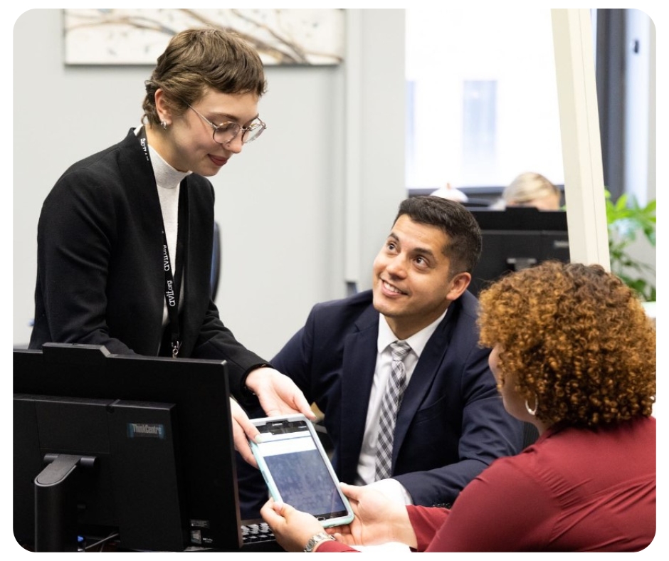 Three people in an office setting. A person with short hair stands at a desk showing a digital tablet to a seated man in a suit and another person with curly hair. Computers and office decor are visible in the background.