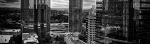 Black and white photo of a cityscape featuring several skyscrapers. The buildings have reflective glass facades, capturing the sky and surrounding structures. A busy urban area is visible below, with roads and greenery in the background.