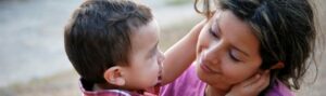 A woman lovingly holds a young child, both gazing into each other's eyes. The child touches the woman's face gently. They are outdoors, and the scene captures a tender moment of affection.