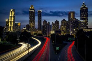 A vibrant nighttime cityscape with tall, illuminated skyscrapers in the background. Bright streaks of red and white lights from moving vehicles create dynamic lines along a highway in the foreground. The sky is dark with some clouds.