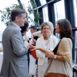 A group of people engaged in conversation at an indoor event with large windows. A man in a gray suit listens to a woman gesturing with her hands. A woman in white stands nearby, observing. Tables with drinks are in the background.