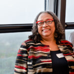 A woman with curly gray hair and red glasses, wearing a striped jacket, is smiling near a window. She has a nametag on her jacket.