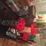 A table filled with various gift bags and boxes in festive holiday wrapping. Some bags have Santa and snowman designs, and others are adorned with red tissue paper. The scene is set in a warmly lit room with a chandelier overhead.