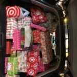 A car trunk filled with wrapped presents in various colorful papers, including red, green, and patterned designs. Some gifts are stacked with visible tags and bows. The trunk is well-lit, indicating holiday preparations.
