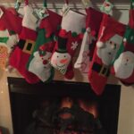 A row of colorful Christmas stockings with festive designs, including snowmen, Santa Claus, and gingerbread men, hangs above a cozy fireplace with glowing embers.
