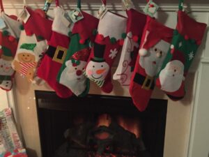 A row of colorful Christmas stockings with festive designs, including snowmen, Santa Claus, and gingerbread men, hangs above a cozy fireplace with glowing embers.