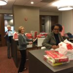 Three women are wrapping gifts in an office setting. One is writing on a card, while the others sort and organize presents. Festive bags and boxes are visible on the tables around them. The atmosphere is busy and cheerful.