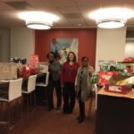 Three people stand smiling in a room filled with wrapped gifts and shopping bags. The room has modern lighting and art on the wall. Tables are used to display the presents.