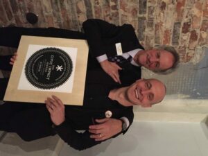 Two men are smiling and standing in front of a brick wall. One is holding a framed plaque that reads 