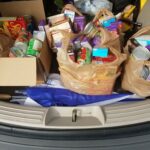 Trunk filled with grocery bags containing canned goods, boxed items, and household supplies. Some bags are plastic while others are cardboard. An umbrella is visible underneath.