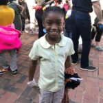A young girl smiling at the camera, standing on a brick path. She wears a yellow shirt and has colorful hair accessories. She holds a piece of clothing in one hand. People in casual clothing are gathered in the background.