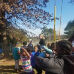Children and adults gather under a tree on a sunny day, looking upward with curiosity. Some are shielding their eyes from the sun. A chain-link fence and trees are visible in the background.