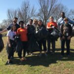 A group of people smiling and standing together outdoors, holding large garbage bags. They appear to be participating in a community cleanup event on a grassy area with bare trees in the background.