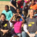 A group of adults and children sit on outdoor steps in the sun. A smiling girl in the center forms a heart shape with her hands. Some wear volunteer T-shirts and gloves. The mood is joyful and relaxed.