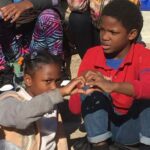 Two children sitting close to each other outdoors, forming a heart shape with their hands. The child on the left is wearing a gray sweatshirt and has colorful hair ties, while the child on the right is in a red shirt and blue jeans. Others are sitting nearby.