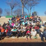 A large group of people, including children and adults, are sitting on outdoor steps. They are smiling and posing for the photo on a sunny day, with bare trees and a clear blue sky in the background. Some hold bags, and the atmosphere is joyful.