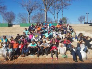 A large group of people, including children and adults, are sitting on outdoor steps. They are smiling and posing for the photo on a sunny day, with bare trees and a clear blue sky in the background. Some hold bags, and the atmosphere is joyful.