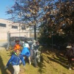 Children in winter clothing gather outdoors near a chain-link fence, under a tree with brown leaves. A brick building and parked cars are visible in the background. The children appear to be enjoying an outdoor activity on a sunny day.