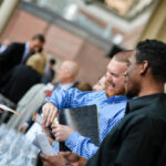 A group of people at a networking event, engaging in conversation near a table with rows of glasses. An excited man in a blue striped shirt holds a folder. The setting is outdoors with a blurred background of other attendees.