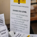 A display with a sign listing sponsors for the "25th Annual Vine Wine Tasting" event under the "Grand Cru Level." Nearby, there are event maps and a wine list in a holder.