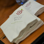 A stack of white napkins with the text "A Divine Event" printed in red and green letters. They are neatly arranged on a wooden surface, with decorative elements visible in the background.