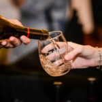 A person pours white wine from a bottle into a glass held by another person. The glass has a logo and text on it. The background is blurred, focusing attention on the wine pouring action.