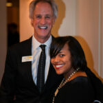A man and woman standing together, both smiling. The man has gray hair and is wearing a suit with a name badge. The woman has black hair and is dressed in black, also wearing a name badge. They appear to be at an indoor event.
