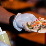 A person wearing gloves holds a taco filled with grilled meat, peppers, onions, and cheese on a small plate. A squeeze bottle of sauce is nearby. The background is softly blurred, focusing on the food.