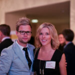 A man in a light blue suit and glasses stands beside a woman in a black dress with a name tag. They are in a room filled with people dressed in formal attire, some holding glasses, suggesting a social event or gathering.