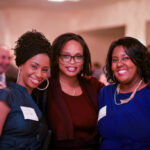 Three women smiling at a formal event, wearing name tags and elegant attire. The background shows other attendees socializing in a warmly lit room.