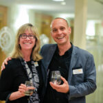 A man and a woman are smiling and standing together indoors. They both hold glasses, possibly of wine, and are wearing name tags. The man is wearing a suit jacket, while the woman is wearing a scarf and a patterned shirt.