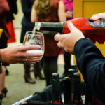 A person in a dark suit pours sparkling wine from a red bottle into a glass held by another individual. The event appears social, with several people in the background, some holding drinks.