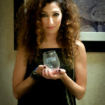 A woman with curly hair holds a glass award with text that reads "Café Latino of the Radio and Music Industry." She is wearing a dark outfit and standing in front of a framed picture with a chandelier.