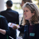 A woman with shoulder-length blonde hair stands at an event, wearing a striped shirt and dark blazer with a name tag. She is engaged in conversation, holding a wine glass. People are mingling in the blurred background.