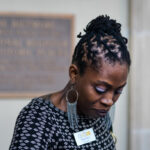 A person with braided hair looks down, wearing a black patterned top and long silver earrings. They have a name badge on their chest. A blurred sign is in the background.