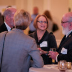 A group of four people, three men and one woman, are engaged in conversation at a networking event. They are dressed in formal attire and wearing name tags. In the foreground, there are candles on a round table.