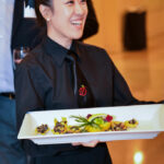 A person in a black uniform is smiling while holding a white rectangular tray with assorted appetizers. They are in a warmly lit room, and a blurred figure in the background holds a glass.