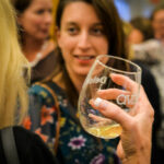 A woman in colorful attire is smiling and talking to another person who's holding a wine glass with blurred people in the background, suggesting a social gathering or event.