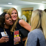 Three women are engaging in conversation at an event. One woman hugs another, who is holding a drink and a card. The third woman, with long blonde hair, listens attentively. People in the background are talking in a well-lit room.