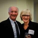An older man and woman smiling in business attire. The woman holds a wine glass and wears a name tag. They are standing in a well-lit indoor setting.