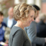 A woman with curly hair is smiling in a semi-formal gathering. She is wearing a gray sweater. The background is blurred, showing people engaged in conversation.