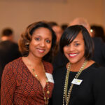 Two women smiling at the camera in a social setting. One wears a patterned red dress, and the other is in a black top with a gold necklace. Blurred people are in the background, suggesting a gathering or event.
